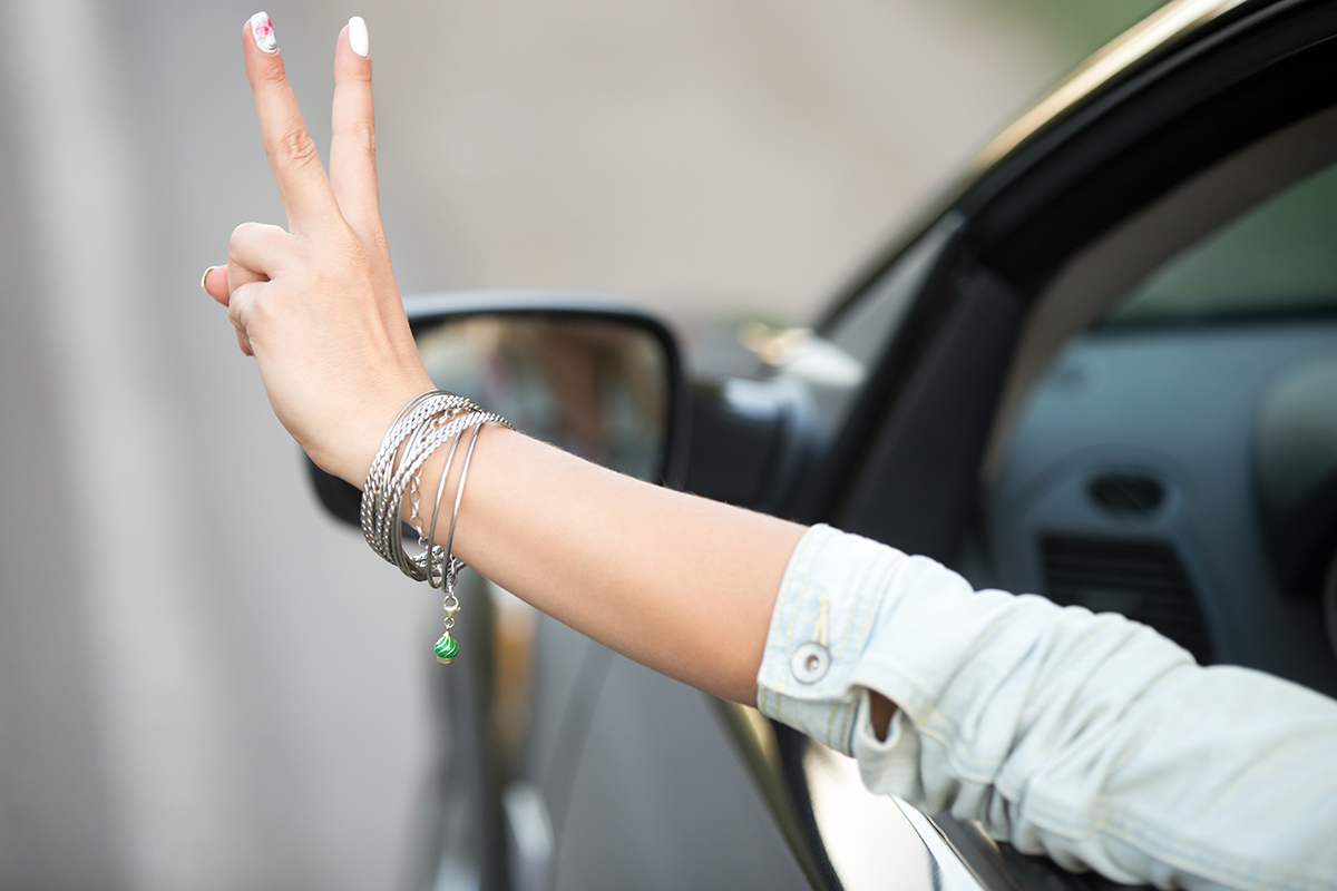Beautiful young woman in casual denim jacket riding modern cabriolet car, showing victory gesture through the window, close up of hand with cute fashion bracelet, back view